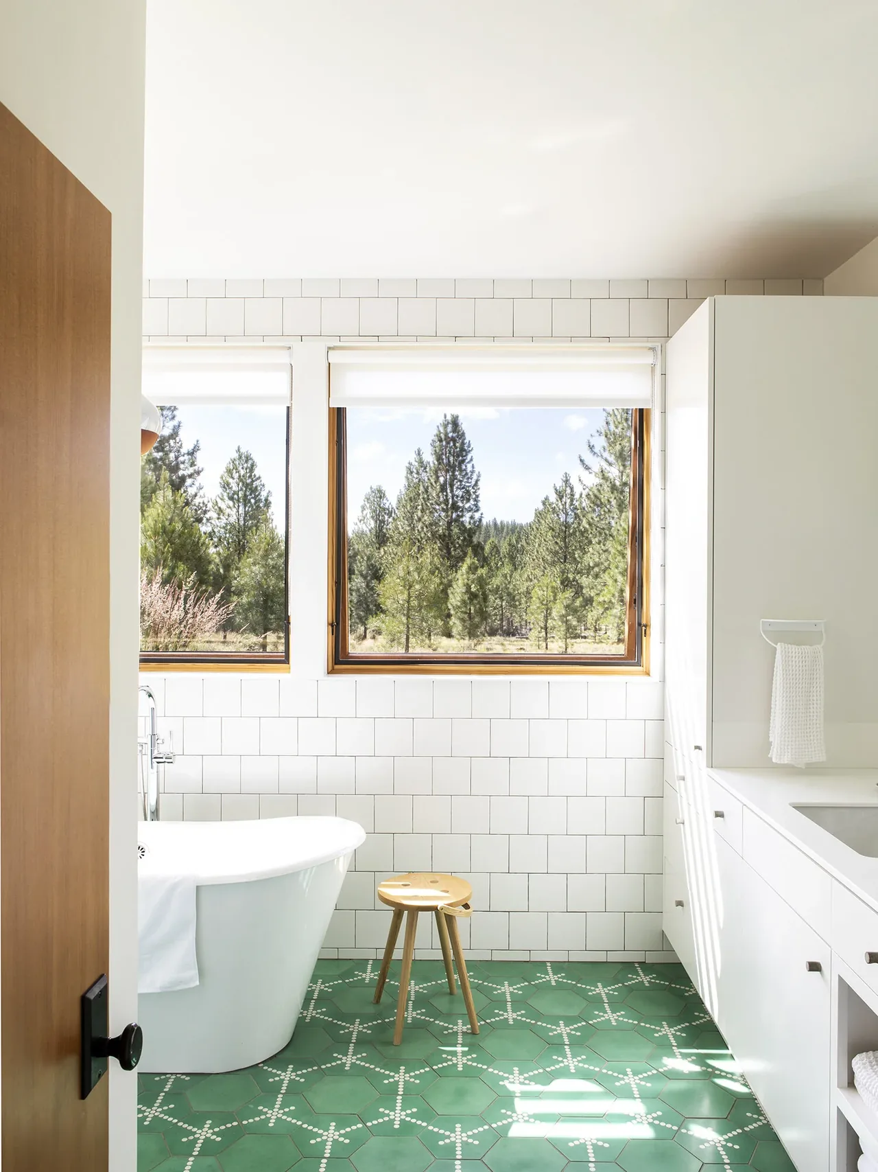 Bathroom with white freestanding tub, green patterned tile floor, white cabinetry, and large windows revealing a pine forest view.