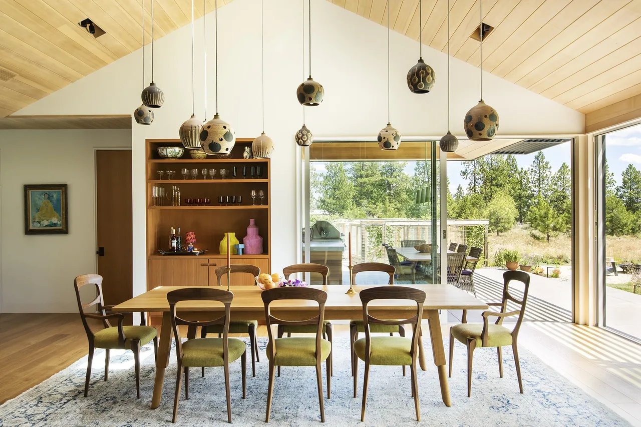 Dining area in BEND HOUSE features a wood table, pendant lights, and floor-to-ceiling sliding glass doors connecting to outdoor patio.