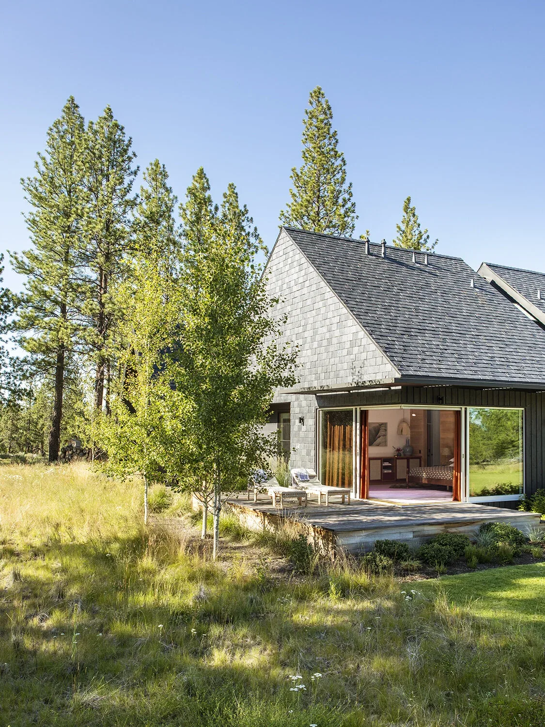 BEND HOUSE features a sharply pitched roof and large sliding glass doors opening to a natural grass landscape with pine trees.