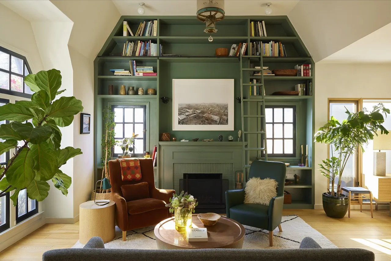 Living room with green built-in bookshelf and fireplace, two armchairs, and large plants by windows.