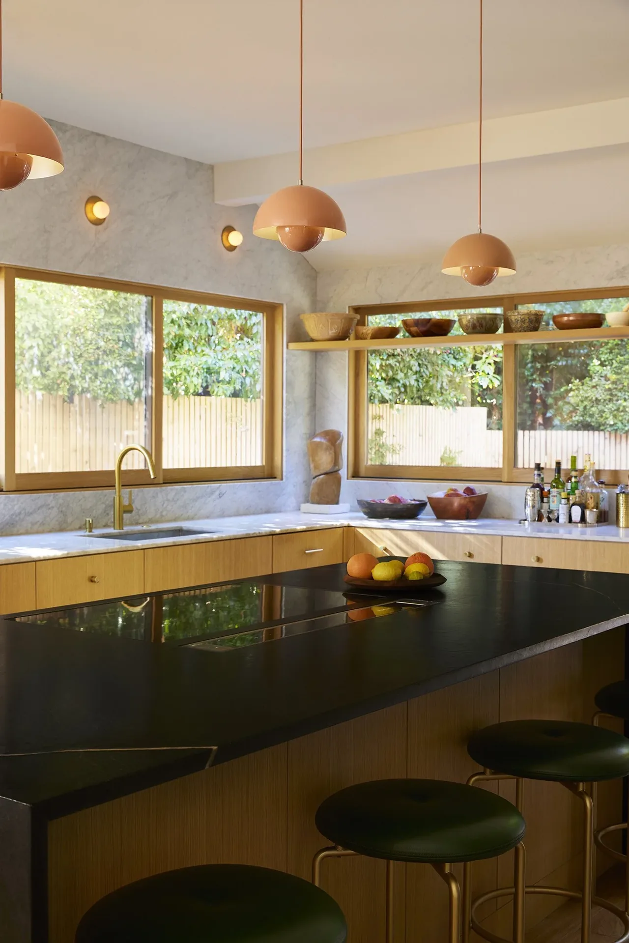 Kitchen featuring a black island countertop, wooden lower cabinets, gold faucet, and terracotta pendant lights in a light-filled space.