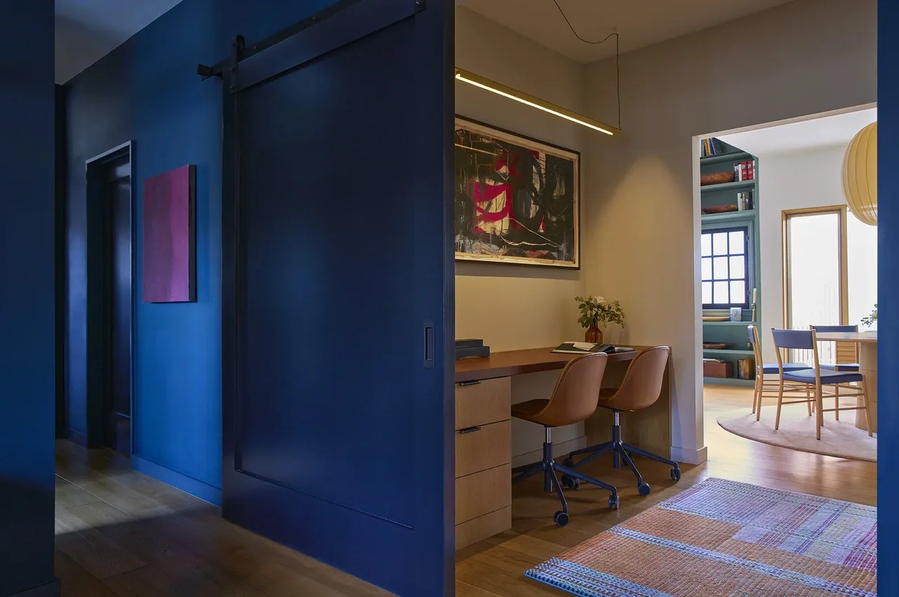 This home office nook features a wooden desk with brown leather chairs and blue sliding barn door, next to a dining area with built-in shelving.