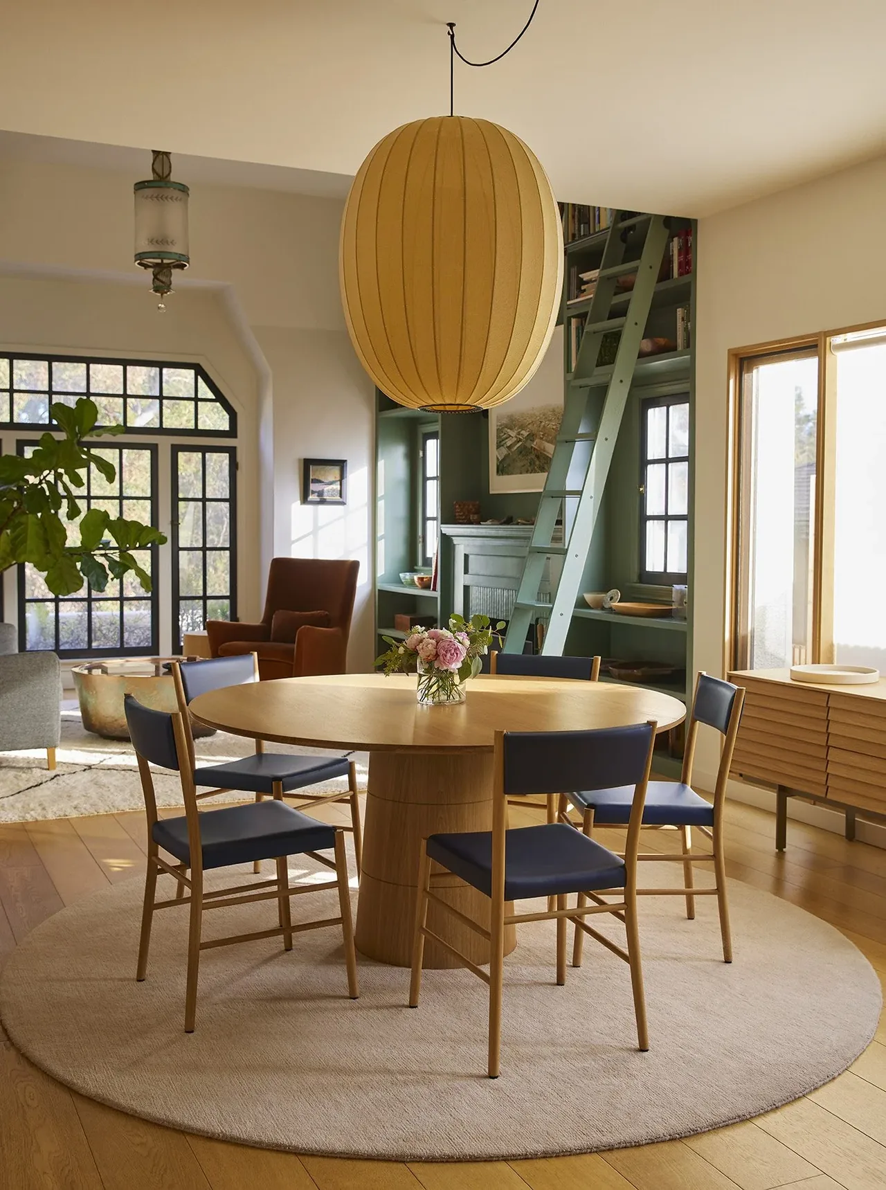 Dining space featuring a light wood round table and dark blue cushioned chairs under a large pendant light.