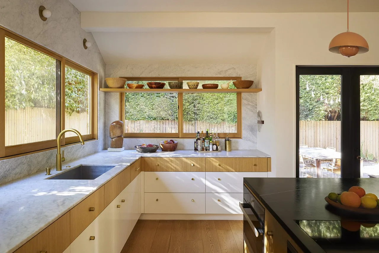 Two tone kitchen features marble countertops, gold faucet, wood framing windows, and a black island with terracotta pendant light.