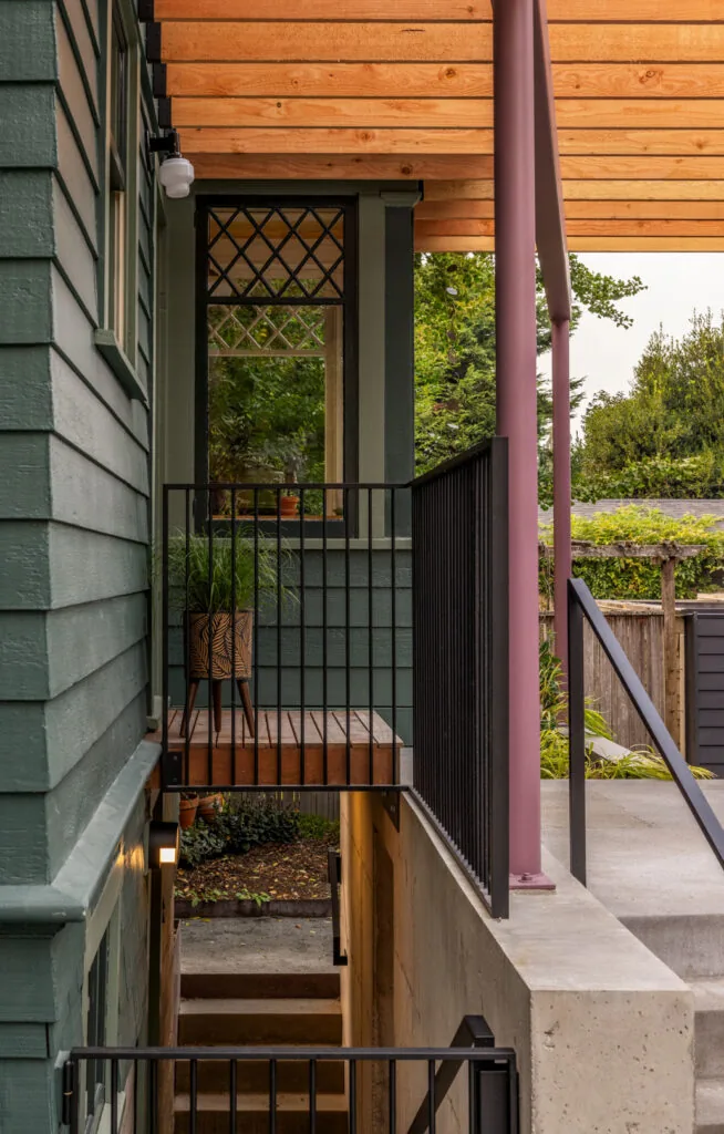 Exterior view of Park Pad showing green siding, a small elevated wooden deck with black railing, and a covered porch with exposed wood roof supported by a pink column.