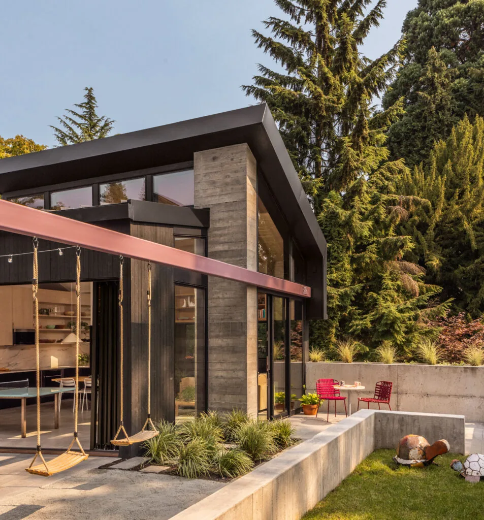 Modern exterior of Park Pad with cantilevered roof, concrete wall, black wood siding, swings, and outdoor seating area.
