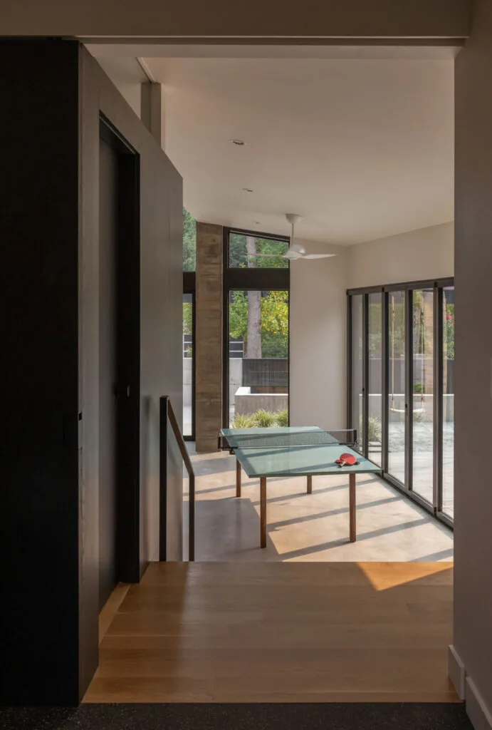 Game room with glass ping pong table and floor-to-ceiling black window frames overlooking outdoor area
