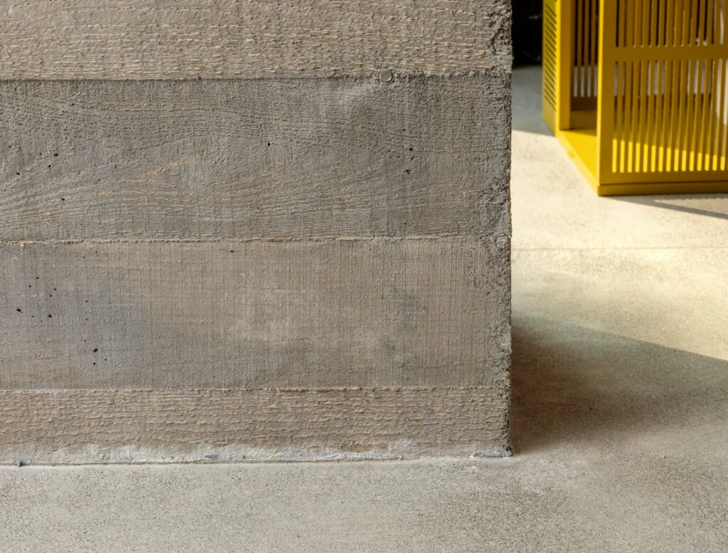 Wood grain textured concrete wall and floor detail with a yellow slatted element visible in the background.