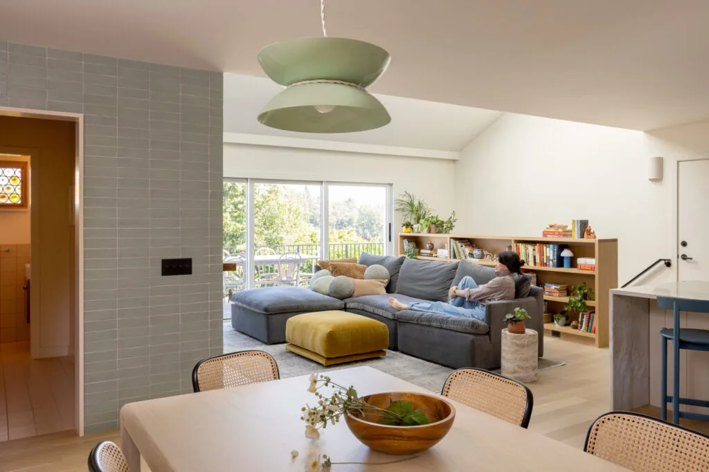 Living room with gray sectional, mustard ottoman, built-in wood shelving, and a view to a balcony through sliding glass doors.
