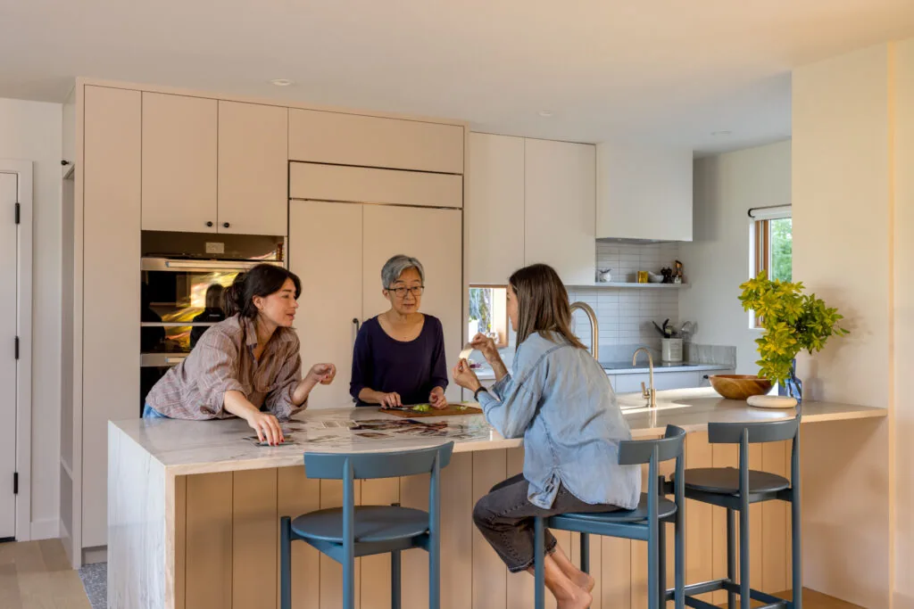 Modern kitchen with large island, blue stools, and built-in appliances where three women converse.