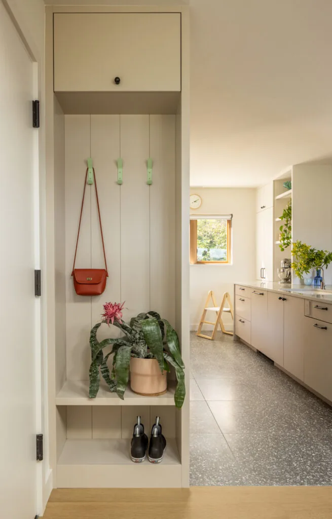 Entry nook with storage hooks, planter, and boots next to a kitchen featuring light cabinetry and terrazzo flooring.