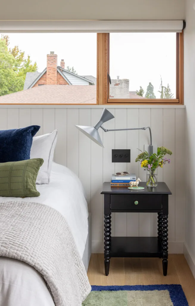Bedroom corner with paneled walls, a black bedside table, and a modern adjustable wall lamp beneath a wood-framed window.