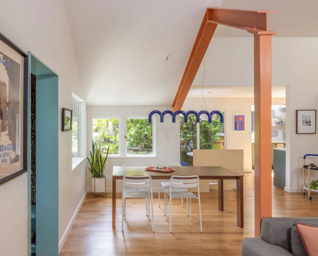 Dining space with a rectangular wooden table, modern white chairs, and exposed orange steel beam structure.
