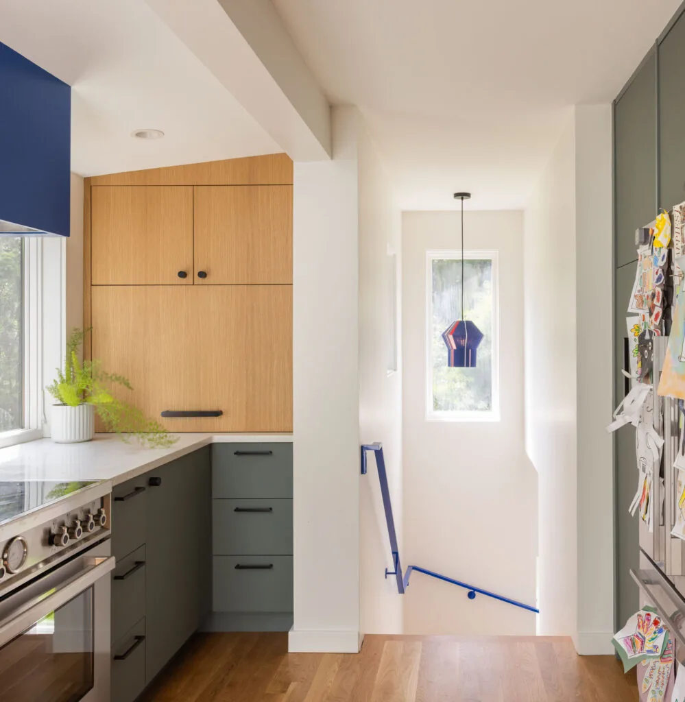 Kitchen with light wood cabinets, blue range hood, hardwood flooring, and a stairway with blue handrail and hanging blue pendant light.