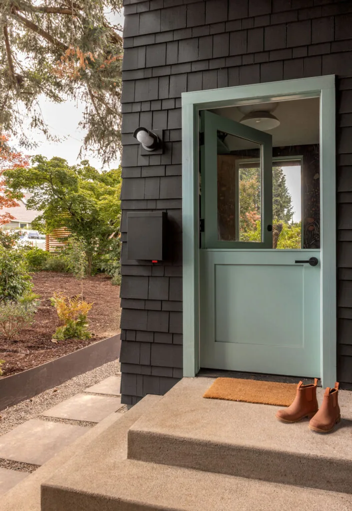 Front entry features a light blue Dutch door set in black shingle siding with concrete steps and a brown doormat.