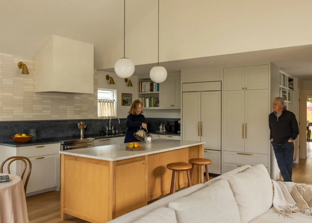 Kitchen features a wooden island with stools, white cabinets with brass handles, and pendant lighting.