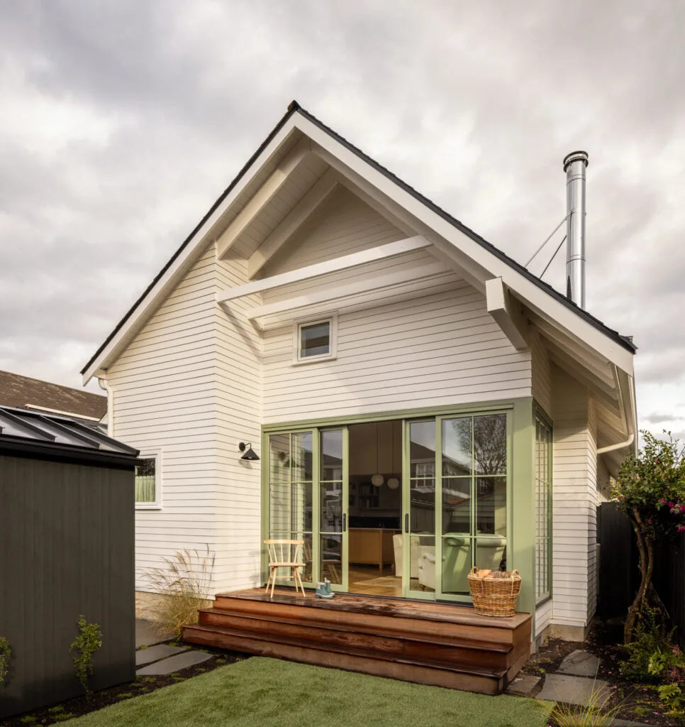 Rad Trad project features white siding, green window frames, a gable roof, and a wooden exterior deck accessed by sliding glass doors.