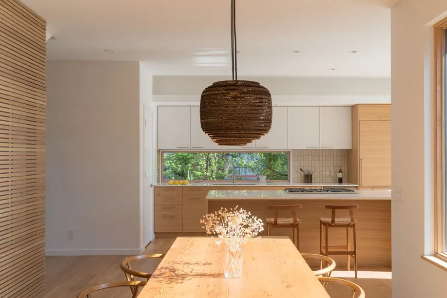 Dining space with a wood table and pendant light facing a kitchen featuring a long horizontal window and wood cabinetry.