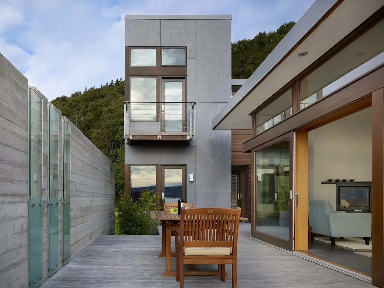 Outdoor dining area on a wood deck overlooking forested hillside with modern concrete and glass elements.