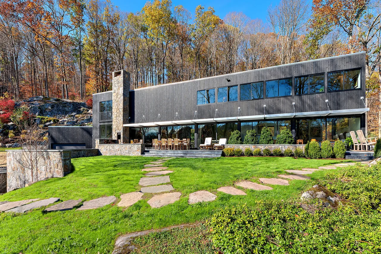 The Stockwell-Rodríguez Residence features dark stained wood siding, a stone chimney, and expansive glazing overlooking a landscaped lawn.