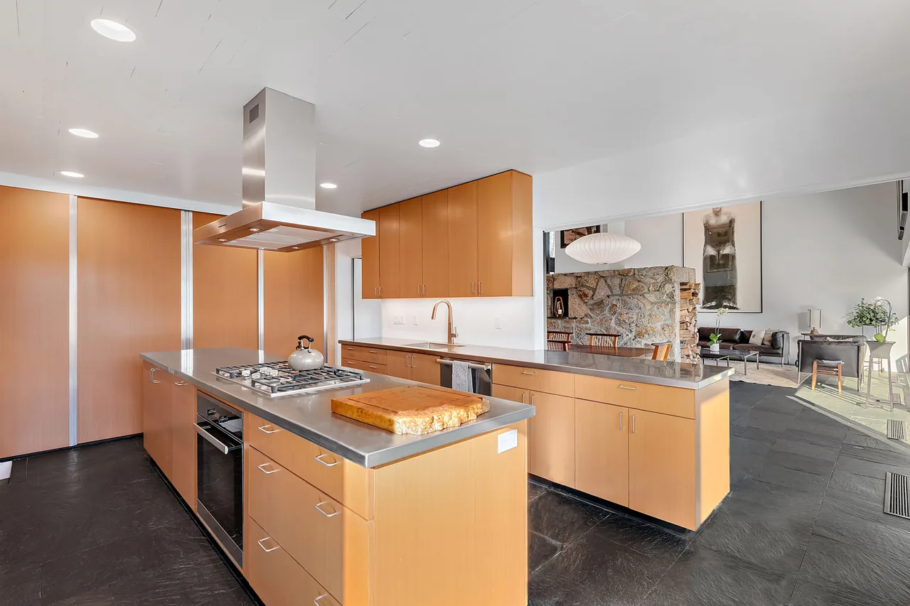 Kitchen of the Stockwell-Rodríguez Residence features wood cabinetry, stainless steel counters, and an island with cooktop under a stainless steel vent hood.