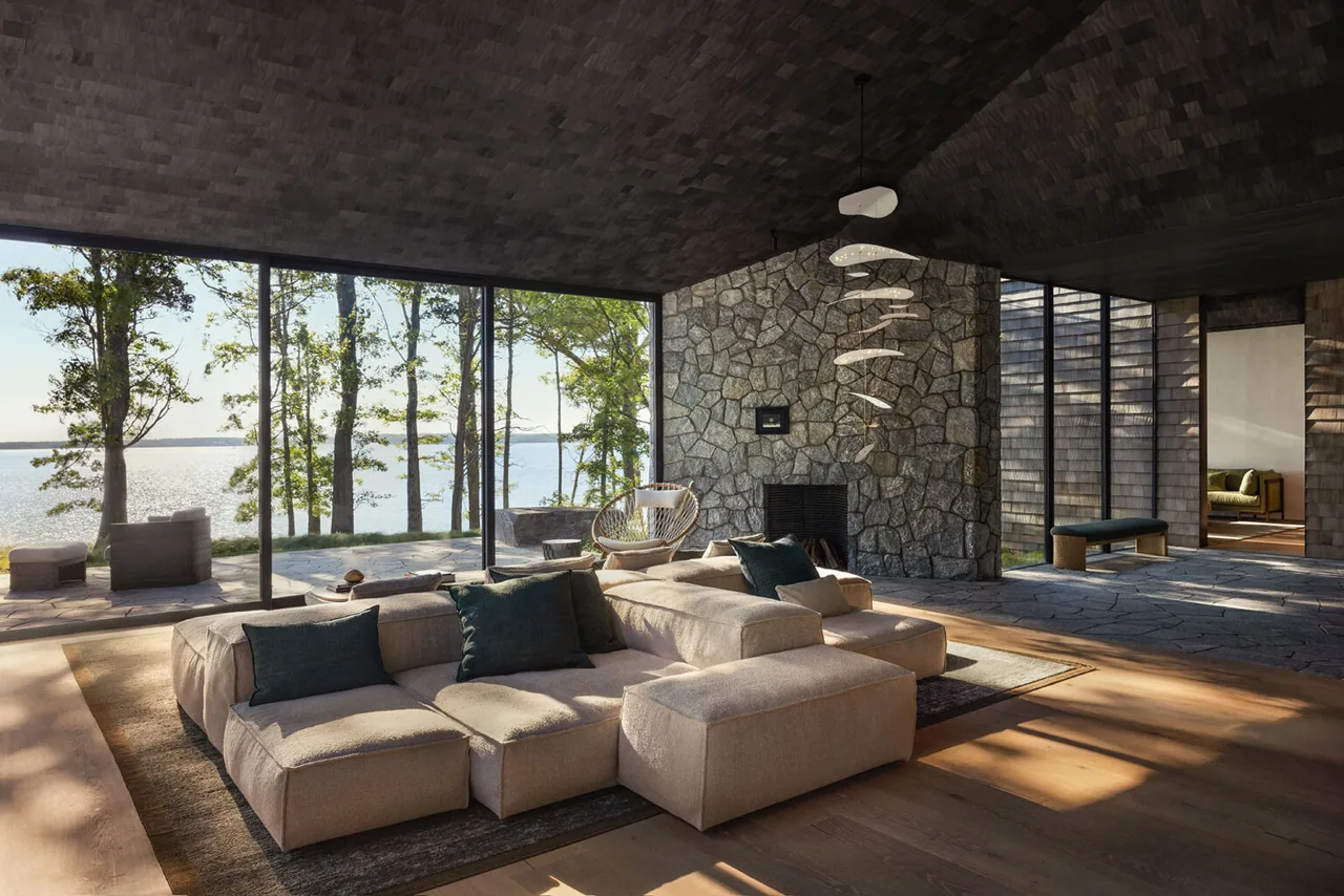 Living room features a stone fireplace, large sectional sofa, and expansive glass windows overlooking the water.