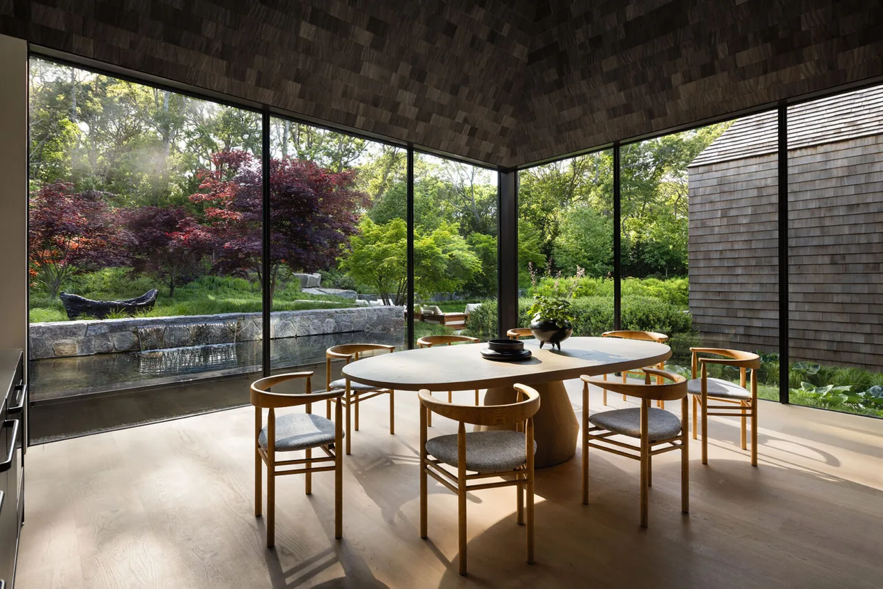 Dining room with a wood table and chairs next to large glass walls showing a landscaped garden outside.