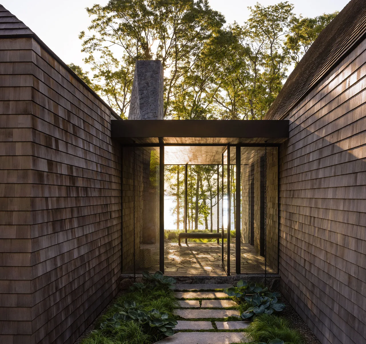 Narrow courtyard features wood shingle siding, stone pathway, glass walls, and a bench overlooking a wooded water view.