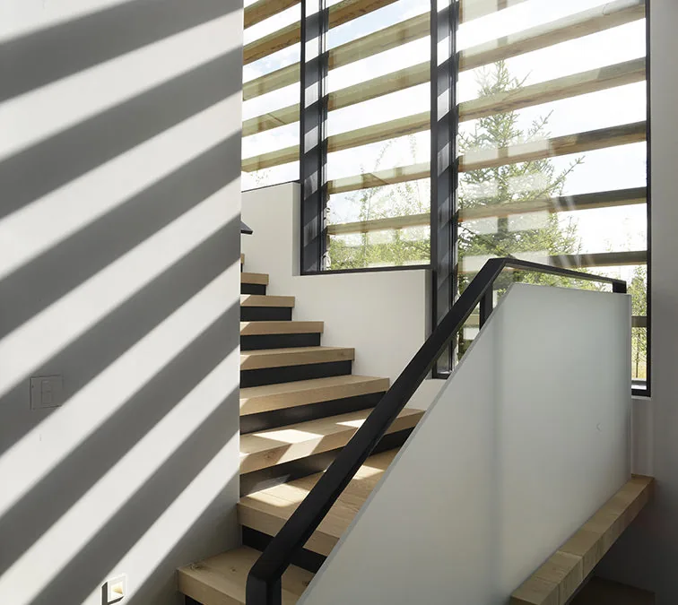 Modern wooden staircase with black railing illuminated by sunlight through large striped windows.