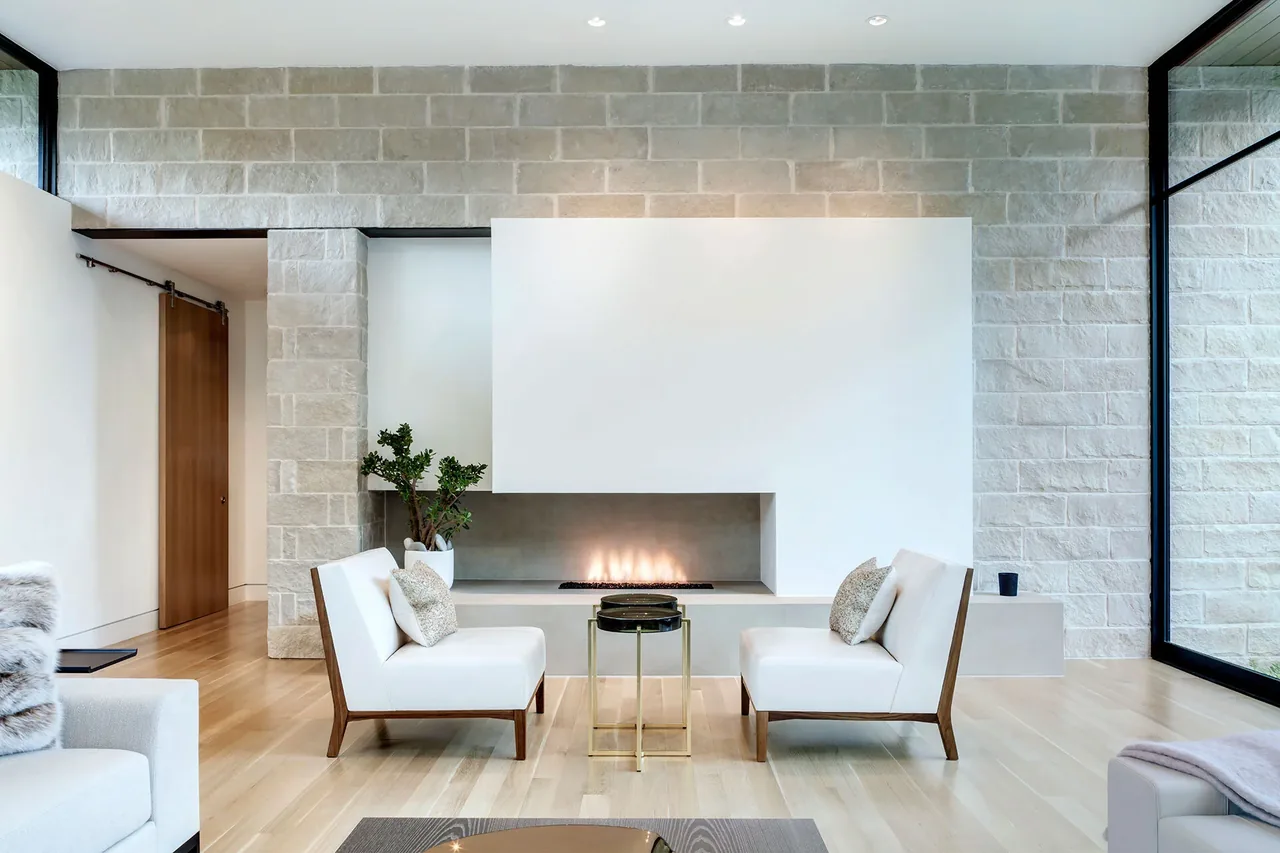 Living room features wood floor, a built-in linear fireplace set into a white wall with stone surround, and white chairs with wood frames.