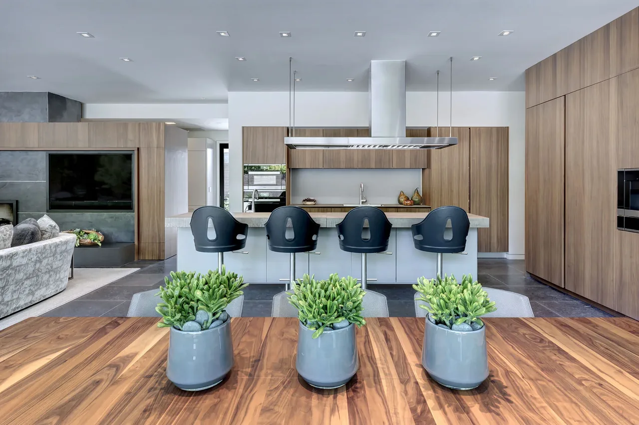 Open kitchen with wood cabinetry, large island with black stools, and gray stone tile floor extending to living area with fireplace surround.