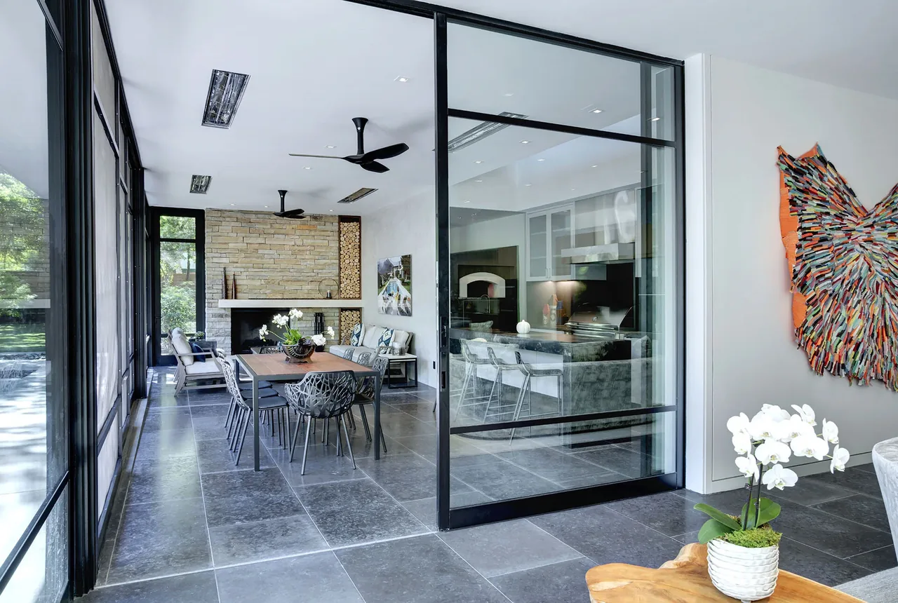 Dining area with wood table and black chairs faces a stone fireplace with stacked wood near large glass walls in Wildwood I project.