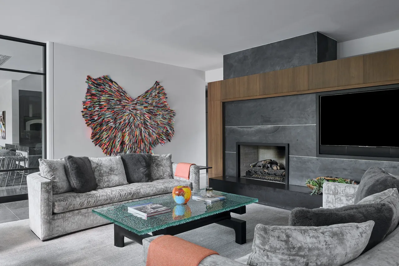 Living room features gray plush sofas, a glass coffee table, colorful wall art, and a modern dark stone fireplace surround with mounted TV.