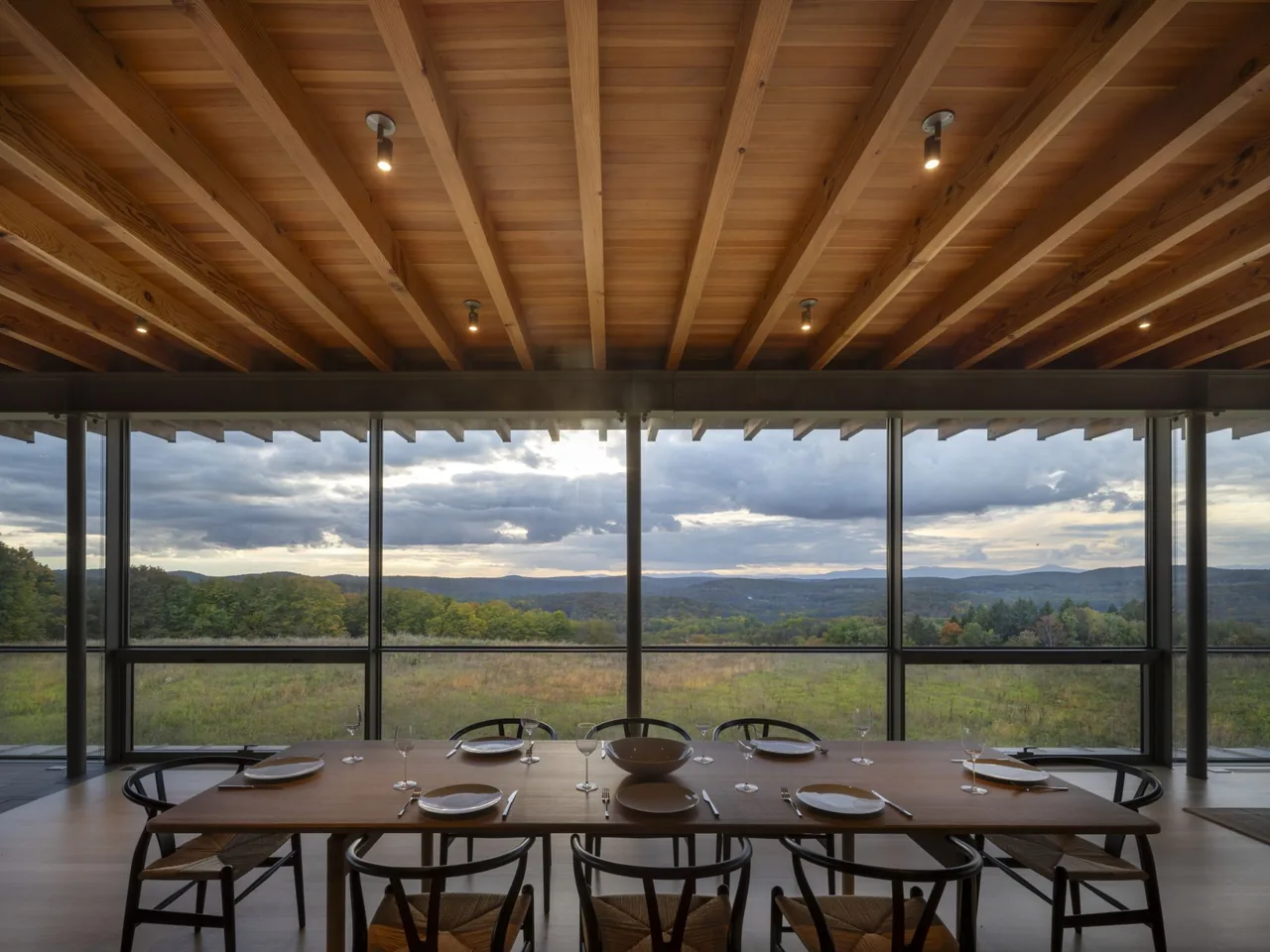 The dining area features a wooden table and chairs beneath an exposed beam ceiling with expansive glass windows showcasing a natural landscape.