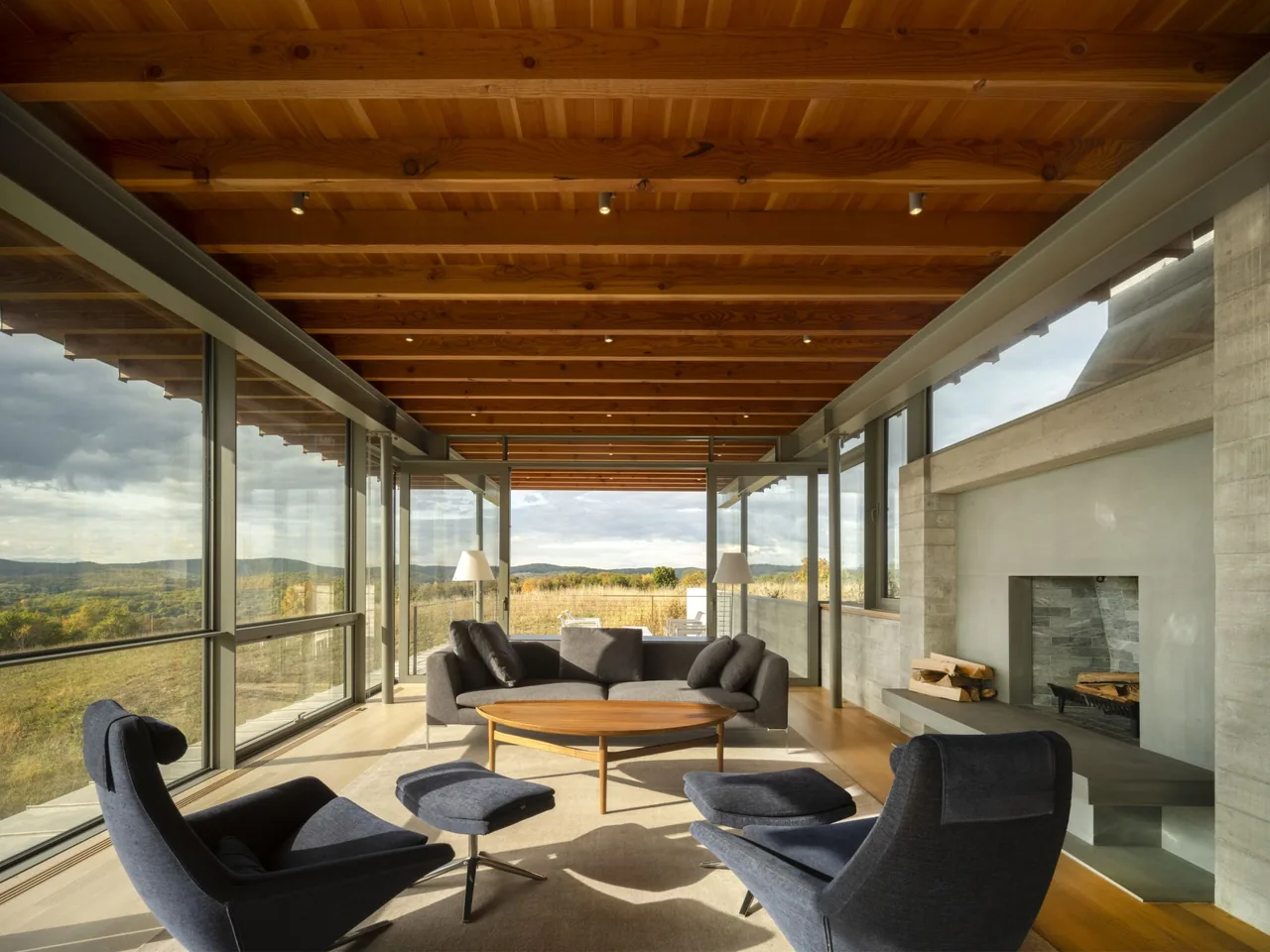 The Gallatin Residence features a living room with a wood beam ceiling, concrete fireplace, and floor-to-ceiling windows showcasing landscape views.