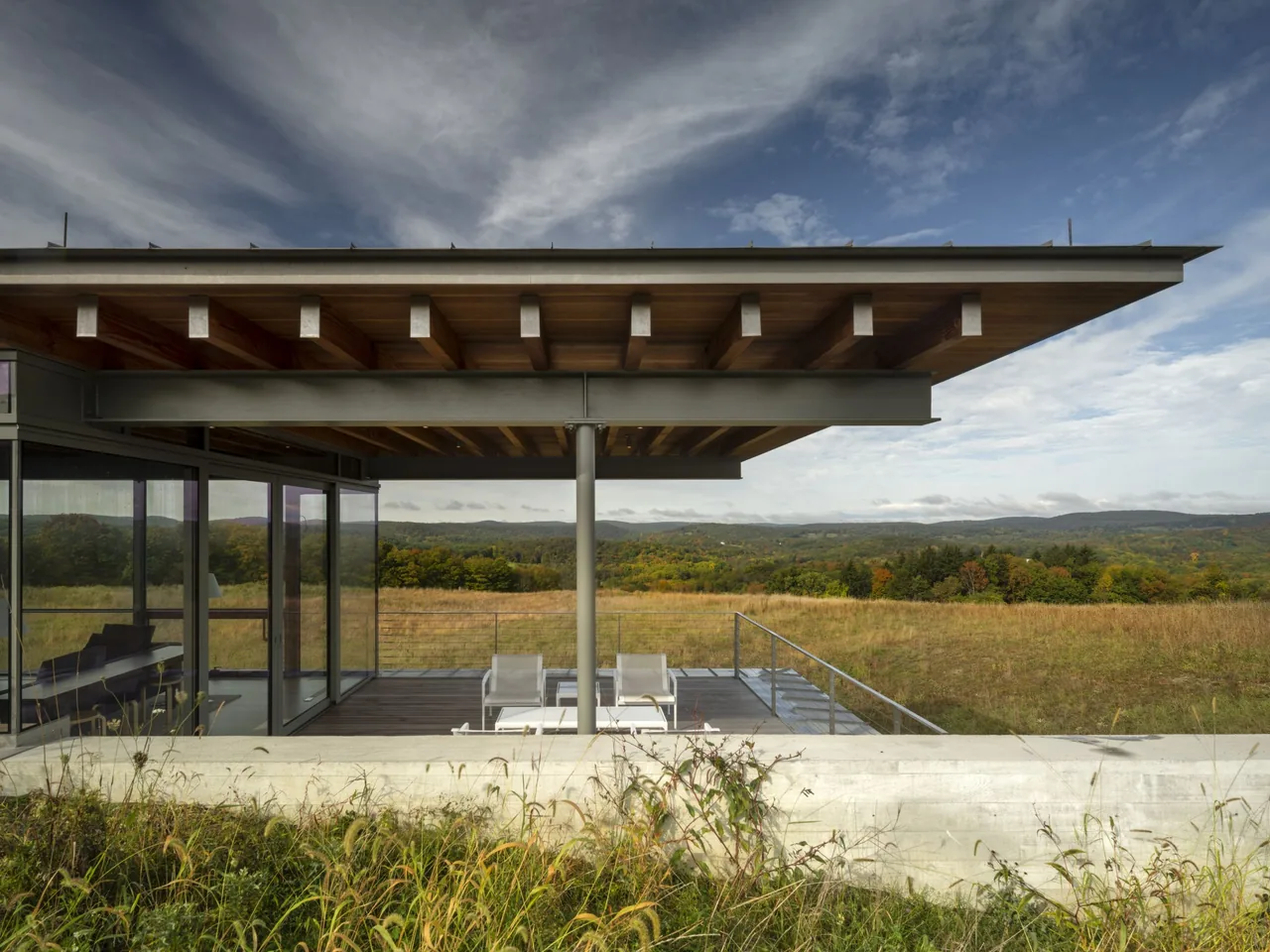 The Gallatin Residence features an overhanging roof with exposed wood beams above an outdoor deck surrounded by glass walls and open views of the landscape.