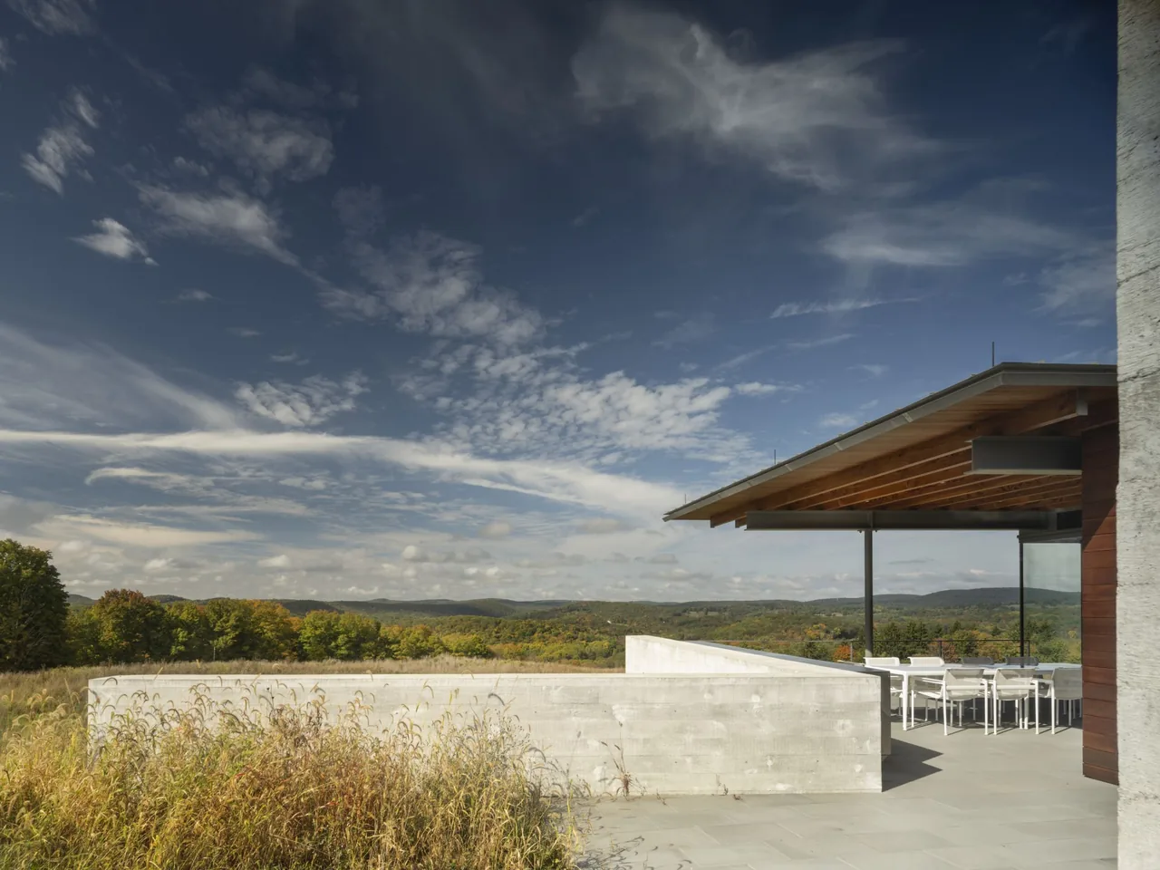 The Gallatin Residence features a covered outdoor dining patio with exposed beams and concrete walls blending into the natural landscape.