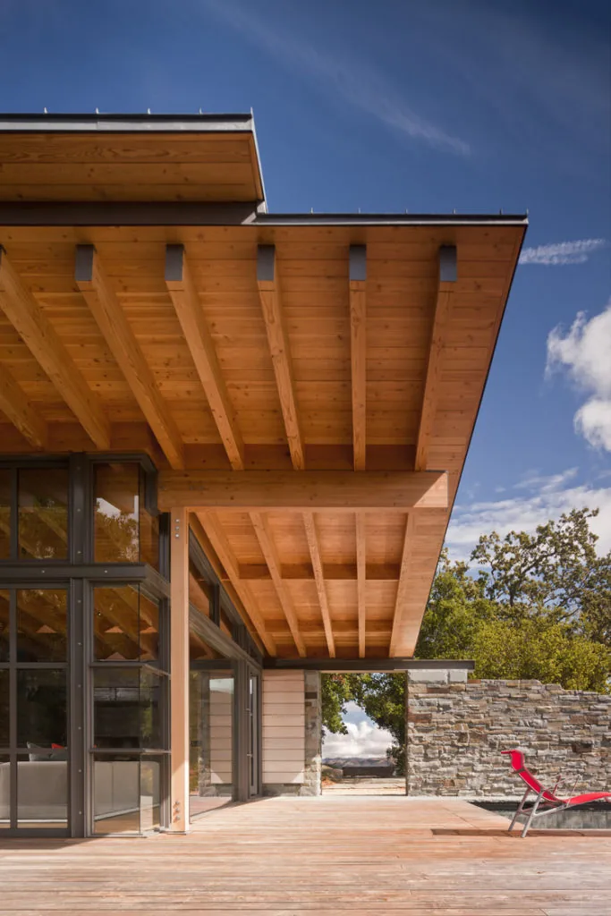 Halls Ridge Knoll Guesthouse features a sloped wood roof overhang, stone retaining wall, and large glass window walls opening to an outdoor seating deck.