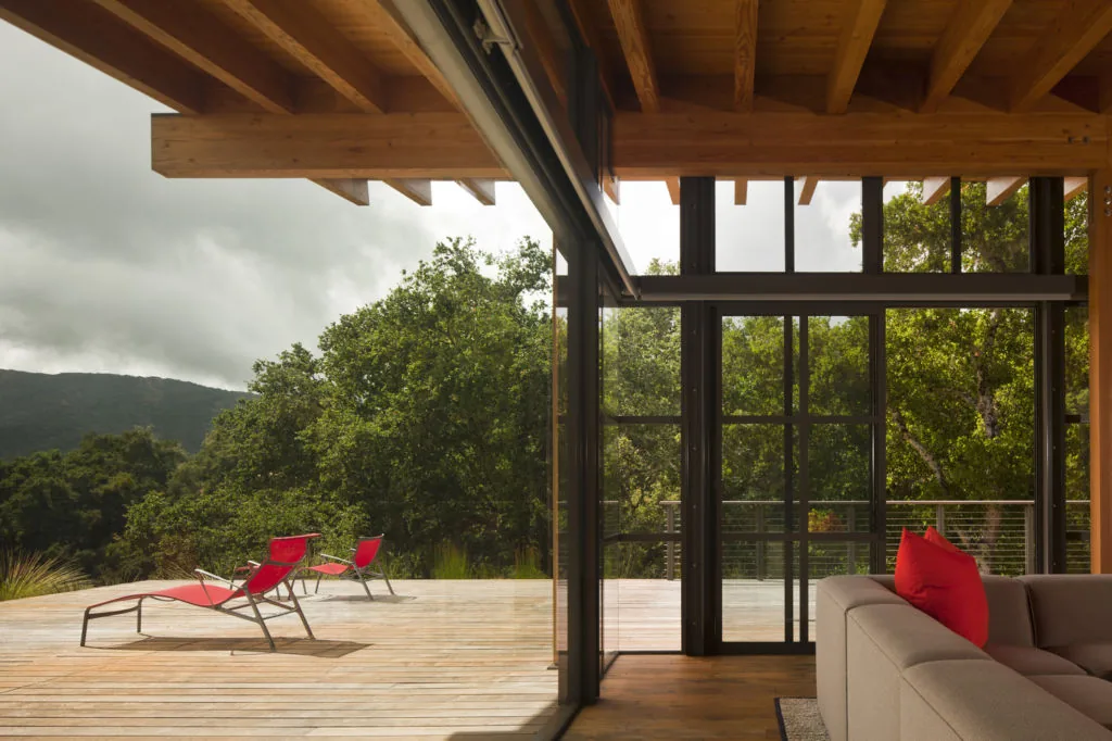 Guesthouse room opening to a wooden deck with red lounge chairs, surrounded by natural landscaping and wood ceiling beams visible inside.