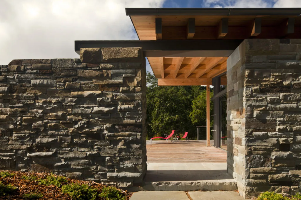 Stone walls frame a wooden deck with red lounge chairs under a sloped wood roof overhang at Halls Ridge Knoll Guesthouse.