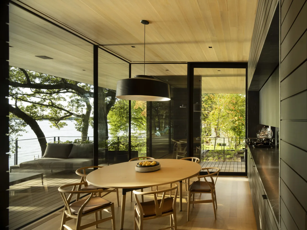 Dining space in Highbank Residence features a round wooden table, floor-to-ceiling glazing, and natural wood ceiling with views of water and trees.