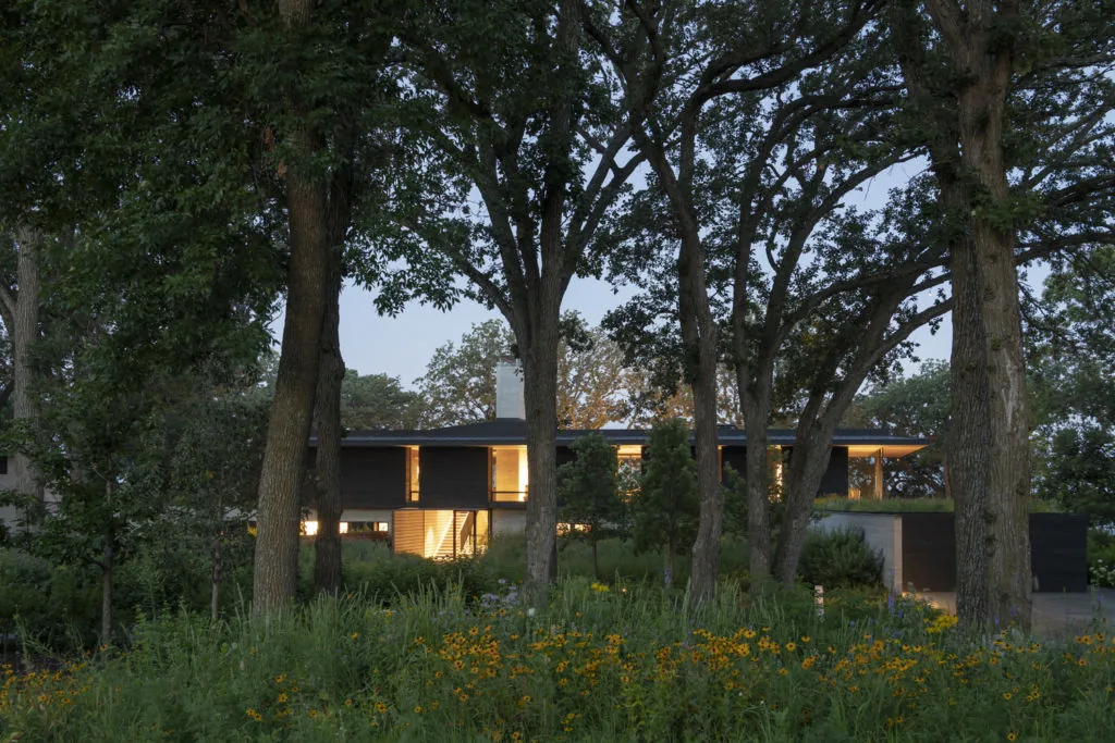 The Highbank Residence features a modern, flat-roofed design illuminated warmly at dusk, set among mature trees and natural landscaping.