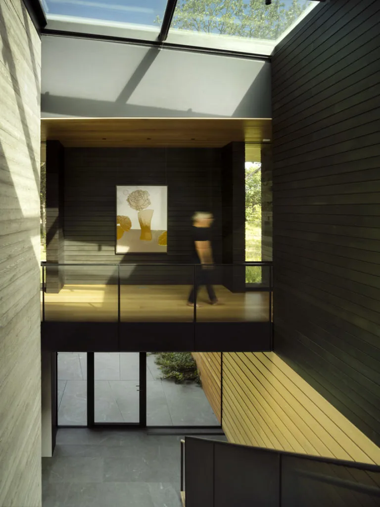 The Highbank Residence interior features a suspended walkway beneath a glass ceiling and integrates black window frames with wood siding and concrete walls.
