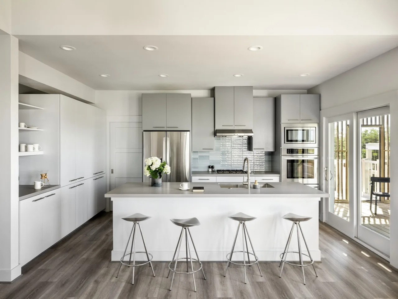 Open modern kitchen features a large island with metal stools, stainless steel refrigerator, double oven, and light gray cabinets.