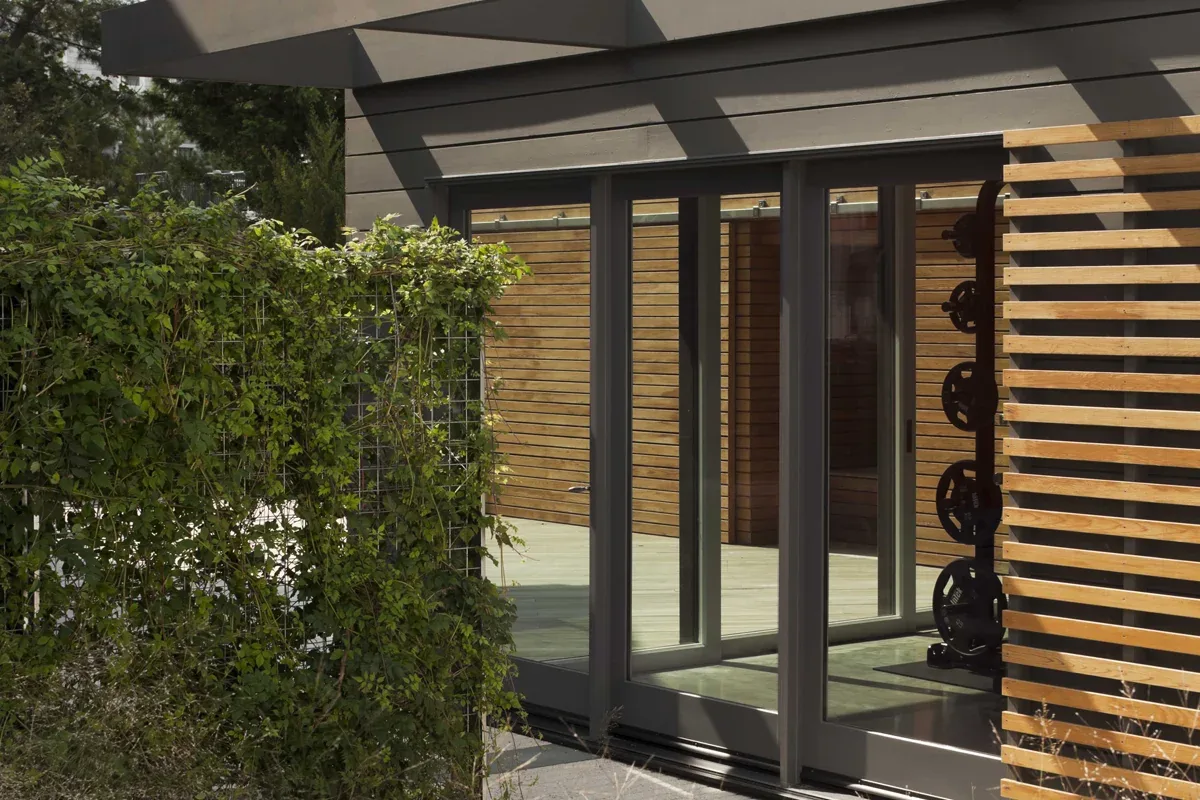 Sliding glass door with black frames opens from the house to a wooden outdoor deck with a privacy screen and greenery.