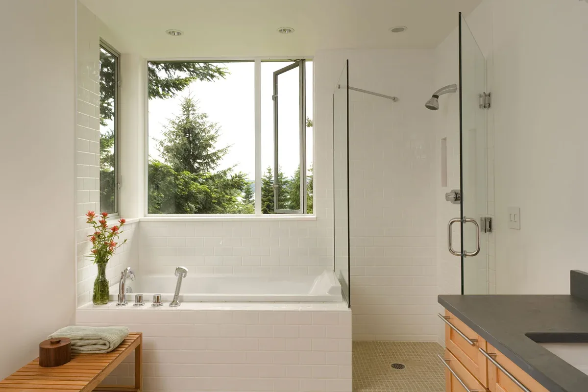 The Geist Residence bathroom features a bathtub beneath a large window and a shower with glass door and tile walls.