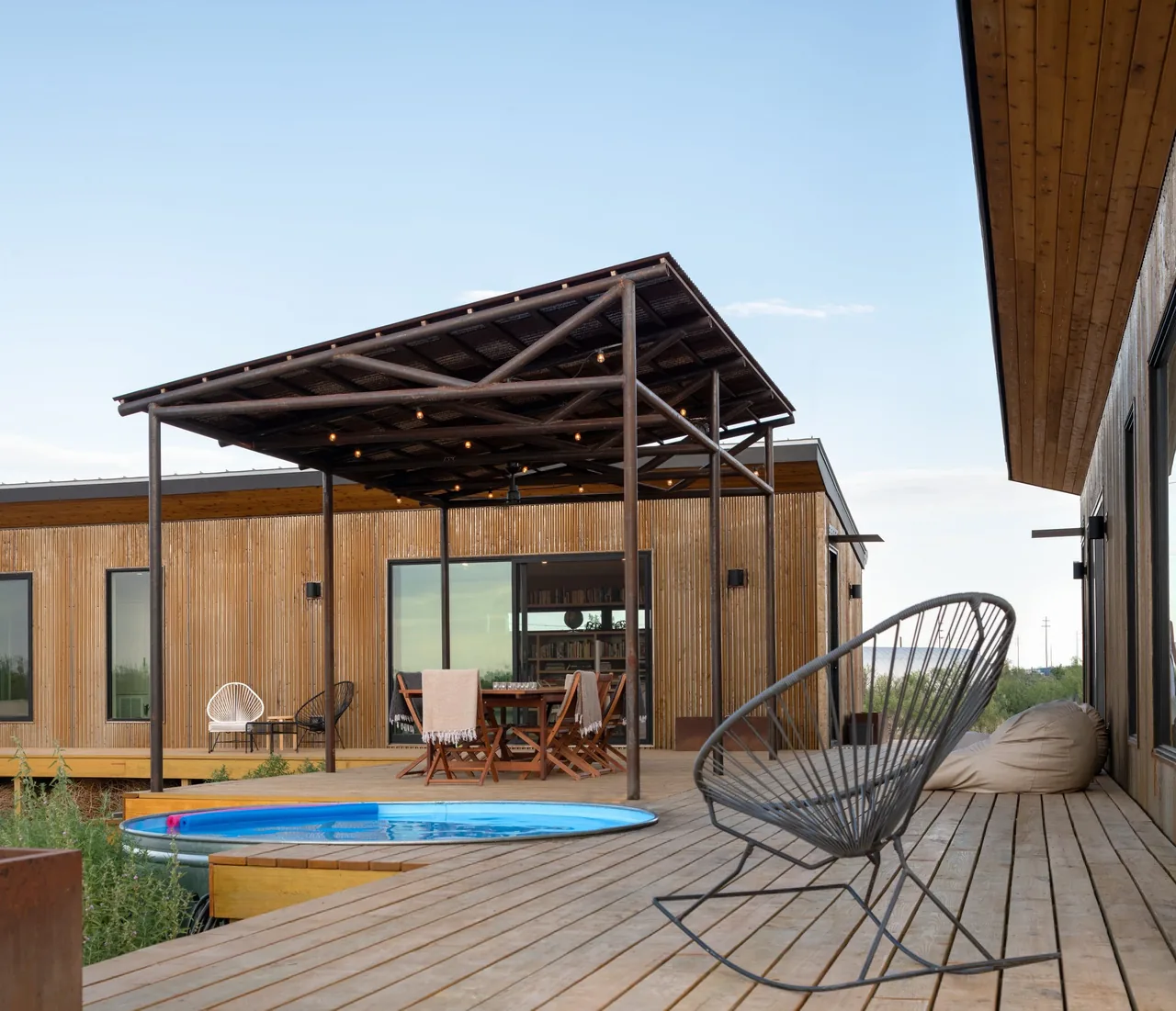 Raised wooden deck with outdoor seating, small round pool, and corrugated siding under a covered patio with string lights.