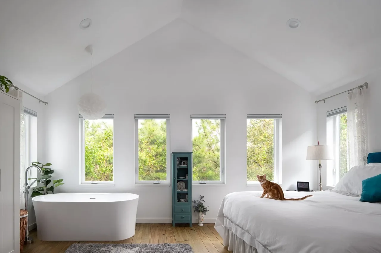 The Saunders House bedroom with vaulted ceiling, large windows, a freestanding tub, and natural wood floor.