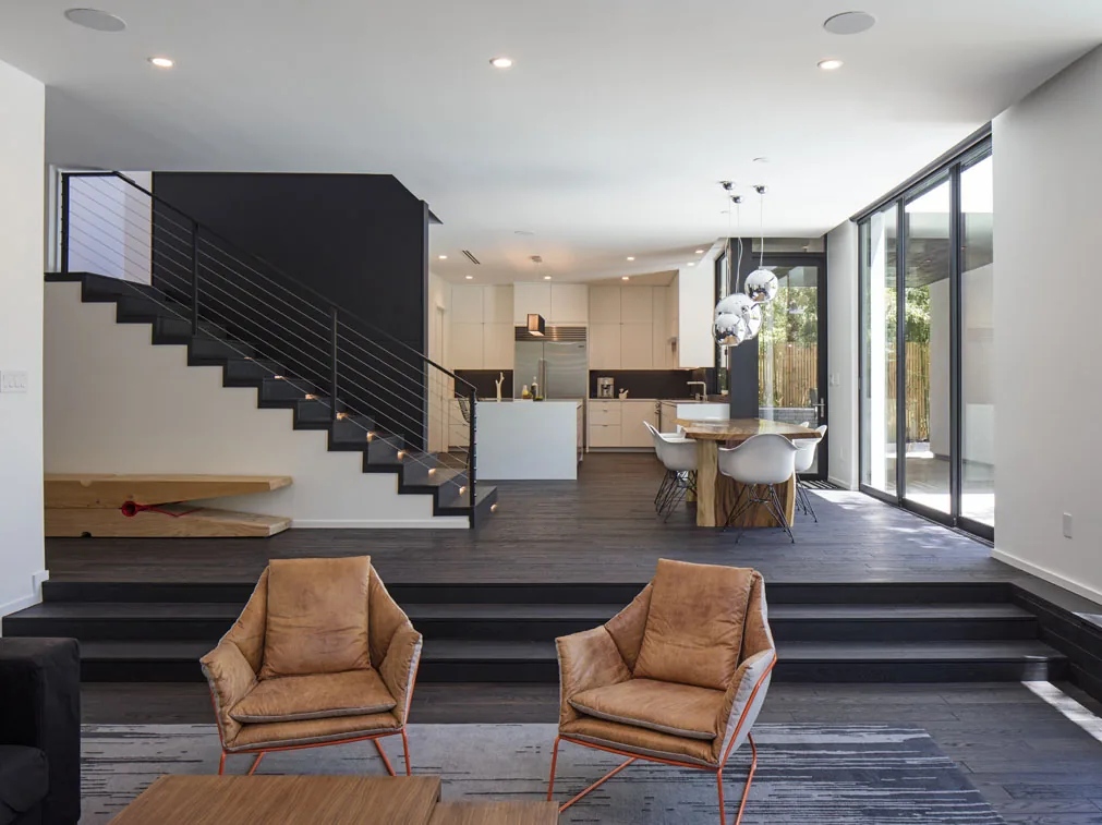 Open living area features black stair with cable railing, white kitchen cabinetry, and a wooden dining table by floor-to-ceiling windows.