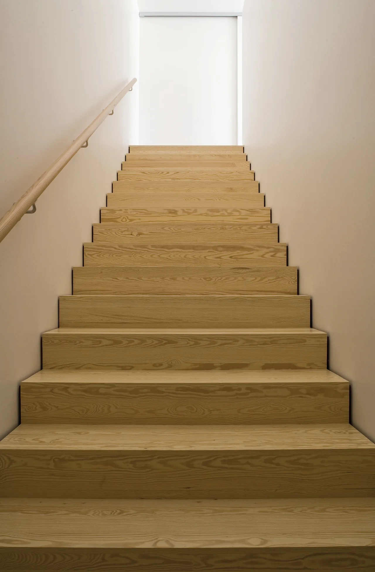 A narrow staircase with light wood steps and a matching handrail ascending between smooth beige walls.